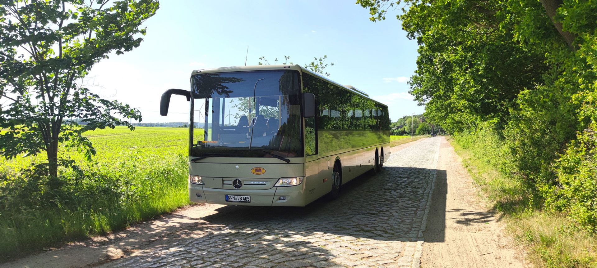 Ein Bus fährt auf einer ländlichen Straße entlang, umgeben von grünen Feldern und Bäumen.